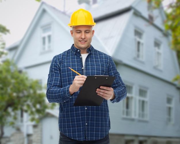 repair, construction, building, people and maintenance concept - smiling male builder or manual worker in helmet with clipboard taking notes over living house background