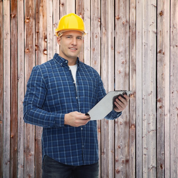修理、建設、建物、人とメンテナンスのコンセプト - 木製のフェンスの背景にクリップボードとヘルメットの男性ビルダーまたは肉体労働者を笑顔