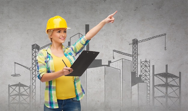 repair, construction and maintenance concept - smiling woman in helmet with construction site on the back with clipboard pointing finger