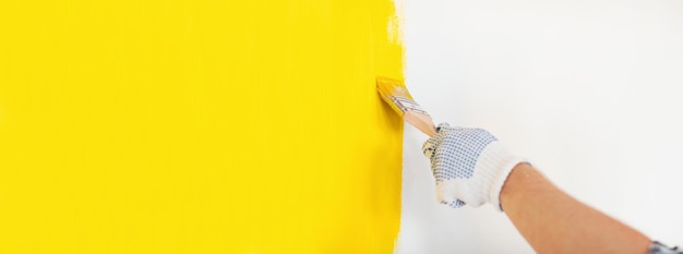 Repair and home renovation concept - close up of male hand in gloves painting a wall with yellow paint