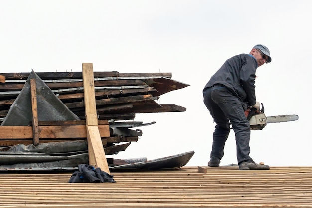 Repair of an old wooden roof Carpenter with tools on the roof against the sky A professional worker in a protective uniform works at the height of a dilapidated house