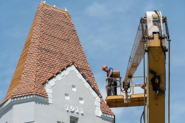 Repair and reconstruction of the old roof made of red tiles of\
a centuryold building the worker is in the basket of a crane and\
works at a height in dangerous conditions roof repair on a sunny\
day