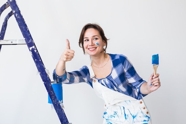 Riparazione, ristrutturazione, lavoratore e concetto di persone - felice giovane donna dipingere il muro a casa, mostrandoci i pollici in su.