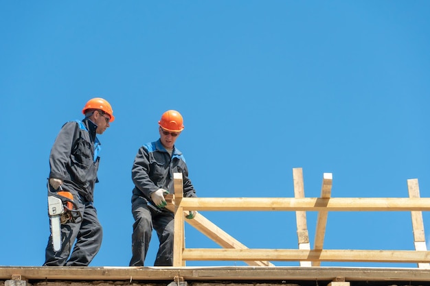 Repair of a wooden roof outdoors against a blue sky background\
two carpenters in special clothes work at a height roofing\
contractors are preparing the roof for the installation of\
insulation