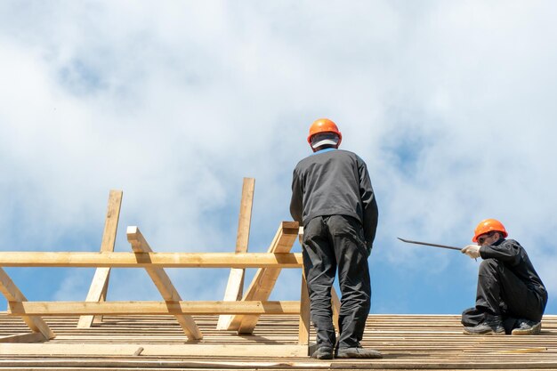 Repair of a wooden roof outdoors against a blue sky background\
two carpenters in special clothes work at a height roofing\
contractors are preparing the roof for the installation of\
insulation