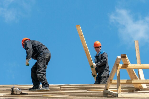 Repair of a wooden roof outdoors against a blue sky background\
two carpenters in special clothes work at a height roofing\
contractors are preparing the roof for the installation of\
insulation