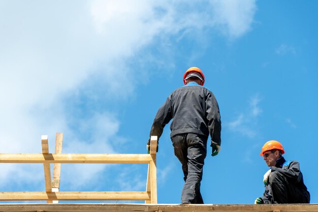 Repair of a wooden roof outdoors against a blue sky background\
two carpenters in special clothes work at a height roofing\
contractors are preparing the roof for the installation of\
insulation