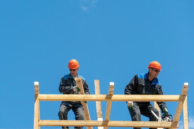 Repair of a wooden roof outdoors against a blue sky background\
two carpenters in special clothes work at a height roofing\
contractors are preparing the roof for the installation of\
insulation