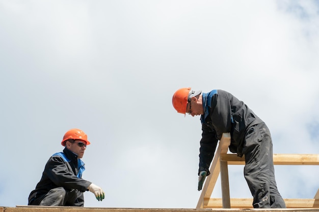 Repair of a wooden roof outdoors against a blue sky background\
two carpenters in special clothes work at a height roofing\
contractors are preparing the roof for the installation of\
insulation