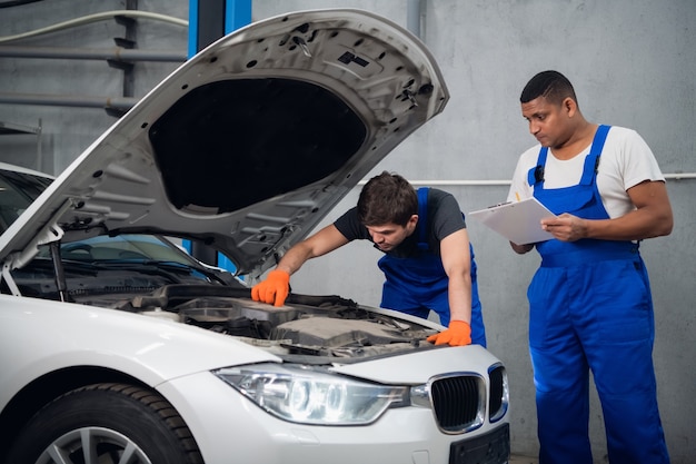 Photo a repairer in blue uniform examines a broken engine of a car. his partner make notes