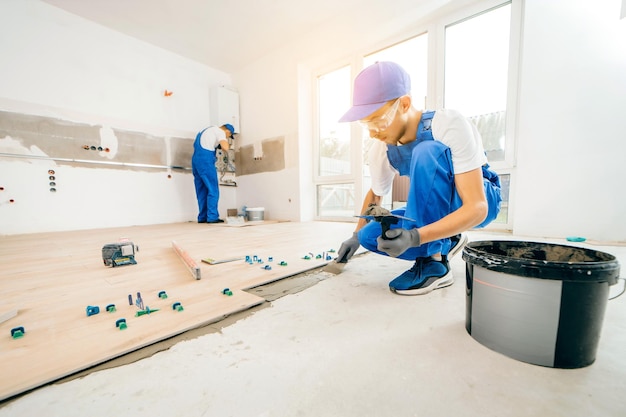 Repairman in grey gloves and uniform laying tiles with tile leveling system on the floor in house