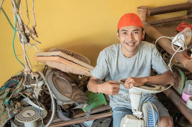 repairman holding the fan cable while sitting around broken items in a service shop