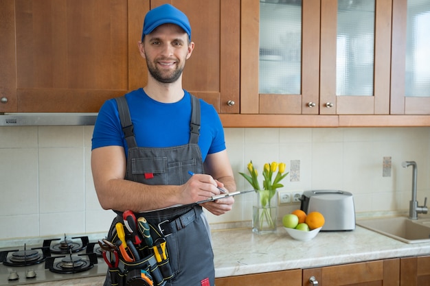 The repairman stands in the kitchen in a blue cap and writes in a tablet