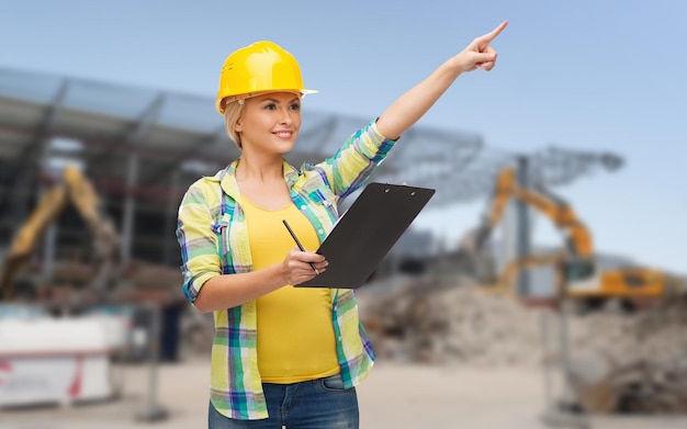 reparatie, bouw, constructie, onderhoud en gebaar concept - lachende vrouw in helm met klembord wijzende vinger