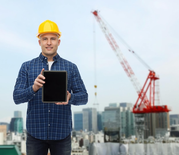 reparatie, bouw, mensen en onderhoudsconcept - glimlachende mannelijke bouwer of handarbeider in helm die het lege scherm van de tabletpc computer over de achtergrond van de stadsbouwwerf toont