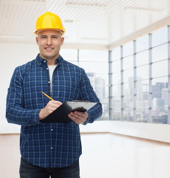 reparatie, constructie, bouw, mensen en onderhoudsconcept - glimlachende mannelijke bouwer of arbeider in helm met klembord die aantekeningen maakt over lege vlakke achtergrond