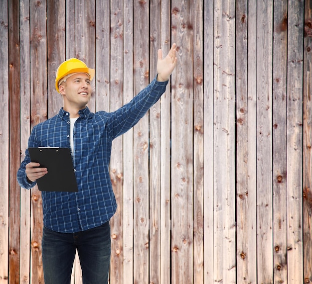 reparatie, constructie, bouw, mensen en onderhoudsconcept - glimlachende mannelijke bouwer of arbeider in helm met klembord wijzende hand over houten hek achtergrond