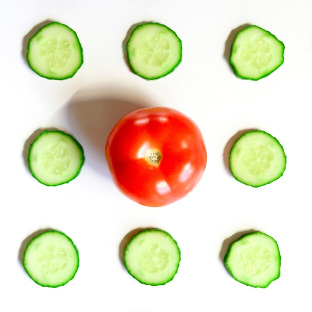 Repeating pattern of sliced semicircles of fresh raw vegetable cucumbers for salad and a whole tomato in the center isolated on a white background flat lay, top view. square