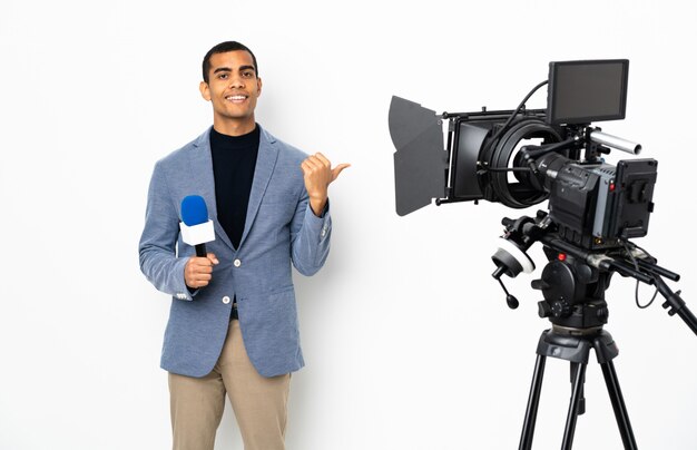 Reporter African American man holding a microphone and reporting news over isolated white wall pointing to the side to present a product