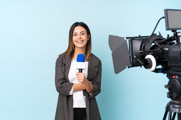 Reporter woman holding a microphone and reporting news over blue wall laughing