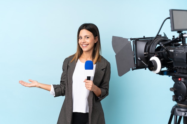 Reporter woman holding a microphone and reporting news over isolated blue wall holding copyspace imaginary on the palm