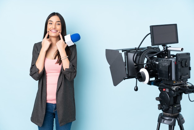 Reporter woman holding a microphone and reporting news isolated on blue wall smiling with a happy and pleasant expression