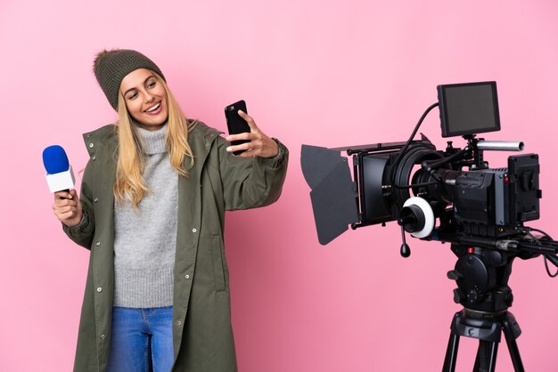 Reporter woman holding a microphone and reporting news over isolated pink background making a selfie