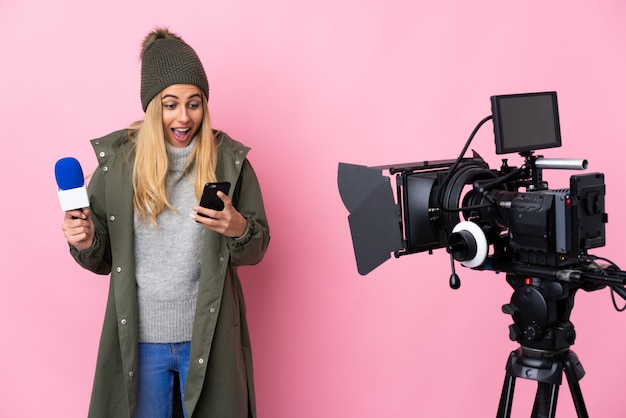 Reporter woman holding a microphone and reporting news over isolated pink surprised and sending a message