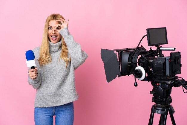 Reporter woman holding a microphone and reporting news over pink wall showing ok sign with fingers