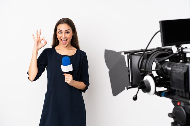 Reporter woman holding a microphone and reporting news on white surprised and showing ok sign