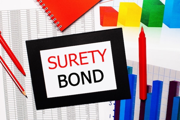 Reports and color charts are on the table. There are also red pens, pencil and paper in a black frame with the words SURETY BOND. View from above