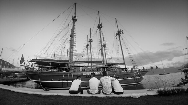 rera view image of people sitting on bench at sea port and looking on moored historical wooden ship