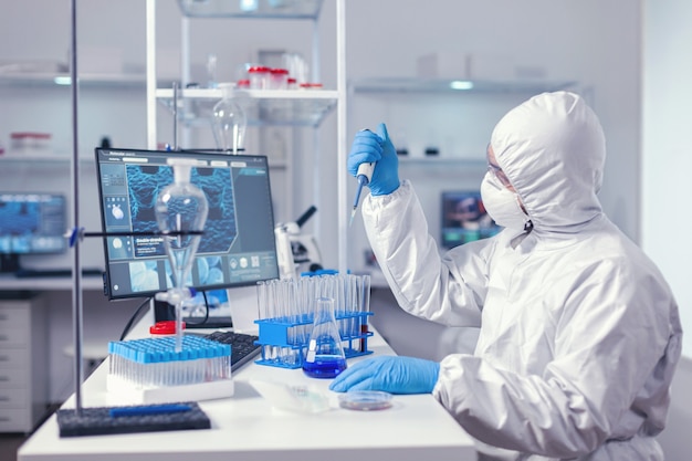 Researcher analyzing sample in automatic pipette working in lab dressed in coverall. Chemist in modern laboratory doing research using dispenser during global epidemic with covid-19.