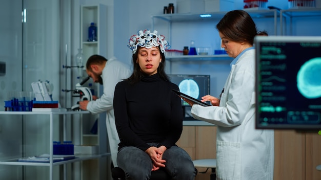 Researcher neurologist doctor asking patient's symptoms taking notes on tablet, adjusting high tech eeg headset. Doctor researcher controlling EEG headset analyzing brain functions and health status