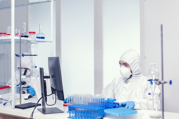 Researcher in sterile suit sitting at workplace in research laboratory for covid19. Medical engineer using computer during global pandemic with coronavirus dressed in coverall.