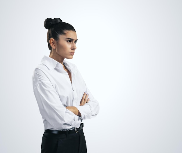 Resentment and unpleasant emotions concept with stressed young woman in white shirt crossing her arms isolated on light grey background close up