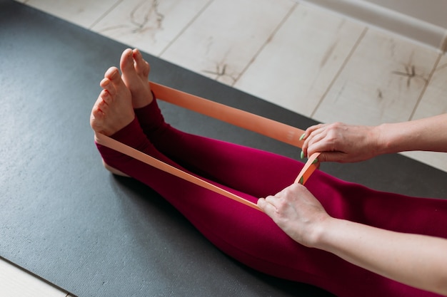Упражнение с эспандером в домашних условиях. Женщина делает тренировку пилатеса, используя эластичный ремень, тянущий руками для тренировки плеч на коврике для йоги в помещении. Концепция спорта и здорового активного образа жизни.