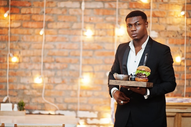 Respectable young african american man in black suit hold tray with double burger against brick wall of restaurant with lights