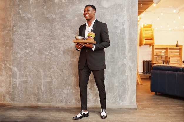 Respectable young african american man in black suit hold tray with double burger against gray wall