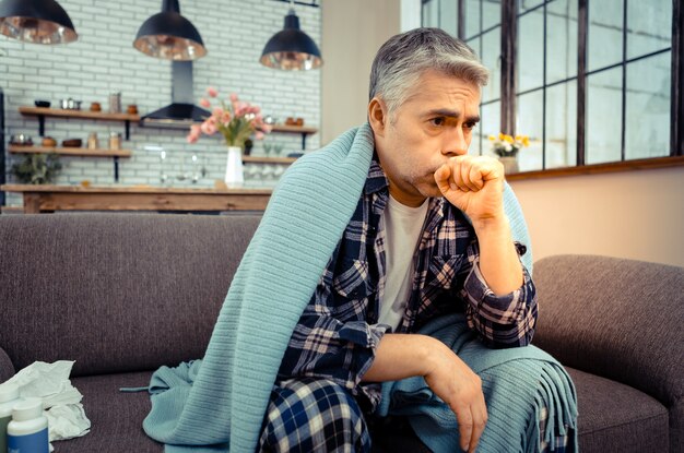 写真 呼吸器疾患。強い咳をしながら自宅のソファに座っている不幸な病人