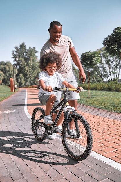 Responsible AfroAmerican dad walking behind a kid riding a bicycle