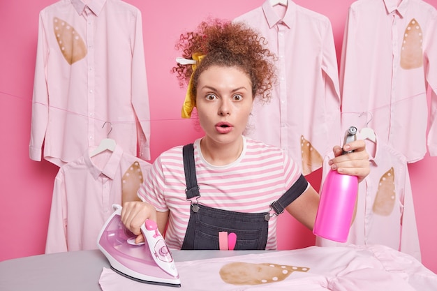 Responsible curly haired woman maid stares bugged eyes busy with housework stands near ironing desk holds water spray bottle and electric iron stands near ironed shirts. Housekeeping concept