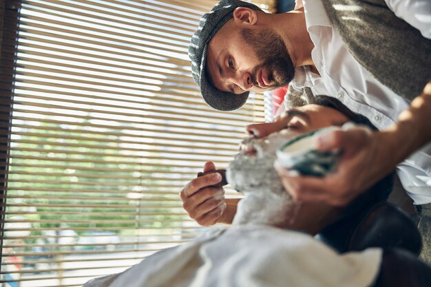 Responsible hair-stylist carefully applying shaving cream on his client facial hair