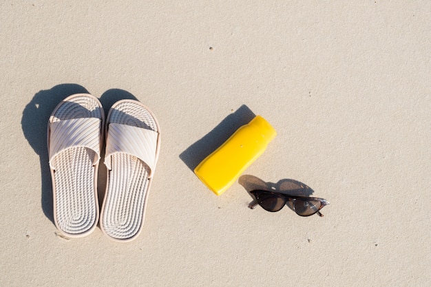 Rest on the beach: slippers, protective cream and sunglasses on clean sand. Top view of acessories for seaside holidays or summer vacation