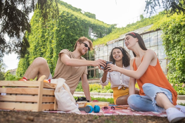 Rest, picnic. Red-haired guy in sunglasses and two long-haired girls holding out hand with drink in great mood at picnic