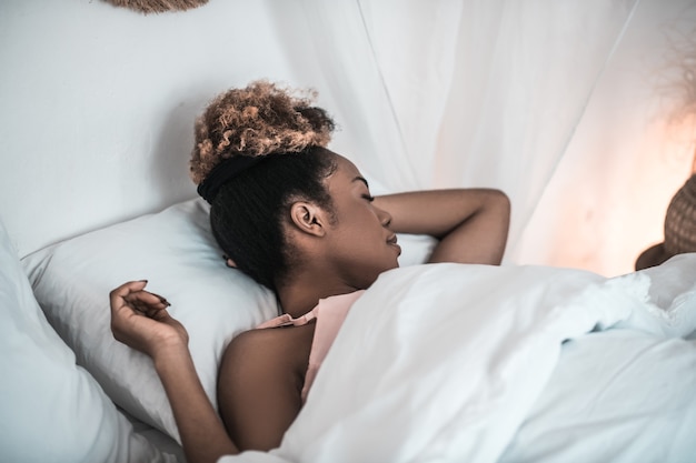 Rest, sleep. Dark skinned young woman sound asleep with hand under her head in white bed in bedroom