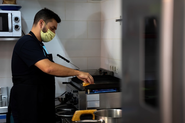 Cuoco di ristorante e bar al lavoro in cucina con la mascherina nella nuova normalità del covid 19