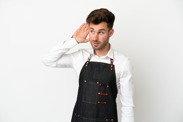 Restaurant caucasian waiter isolated on white background listening to something by putting hand on the ear