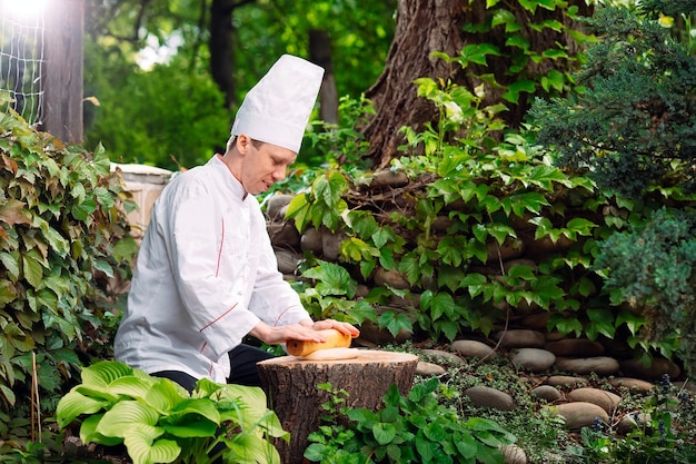 A restaurant chef in uniform rolls out dough on a stump in the woods.