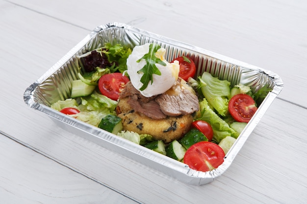 Restaurant food delivery in foil box on white wood table. Poached egg closeup on veil steak medium rare with fresh vegetable salad and couscous cushion. Dish take away closeup, healthy meal.